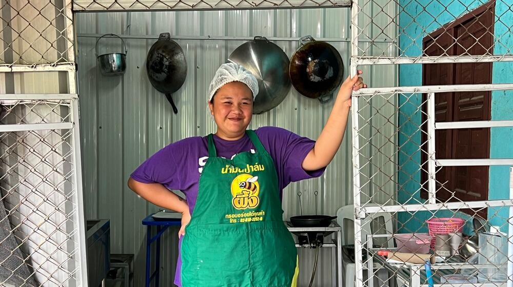 A woman wearing a green apron stands in a kitchen where Dok Jok is prepared.