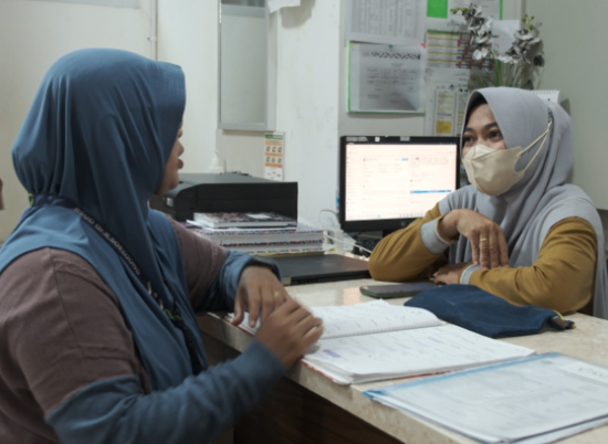 Dos mujeres con las cabezas cubiertas conversan en un consultorio médico con una computadora en el fondo y archivos abiertos frente a ellas