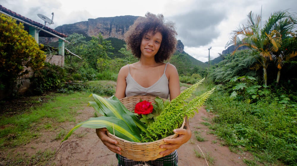 A woman smiles and holds a basket of flowers and plants. 