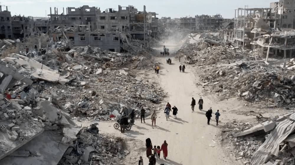  صورة من أعلى لمبانٍ مدمرة بالكامل وأنقاض، مع عدد قليل من  الناس يسيرون على طريق ترابي